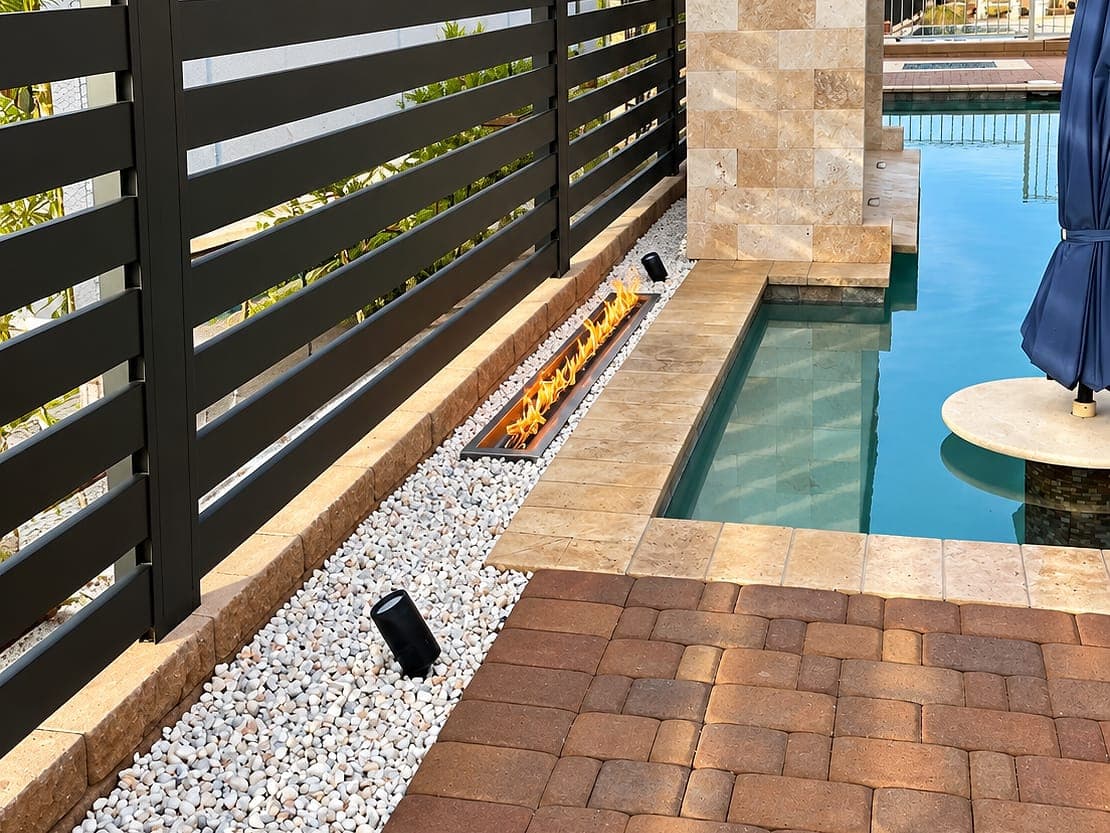 Horizontal aluminum slat privacy fence along a narrow pool with a fire feature and stone water wall.