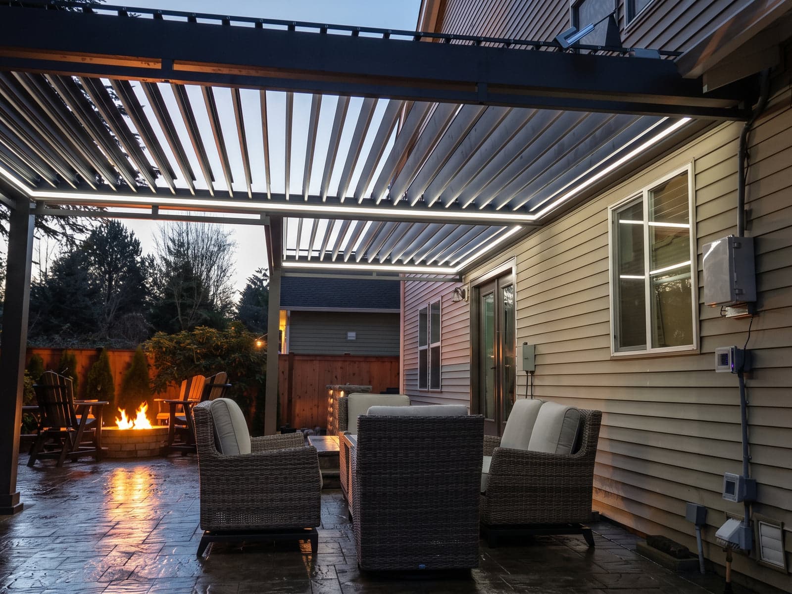 Louvered roof over a patio lounge at dusk, outlined with white LED strip lights, with a fire pit in the yard.