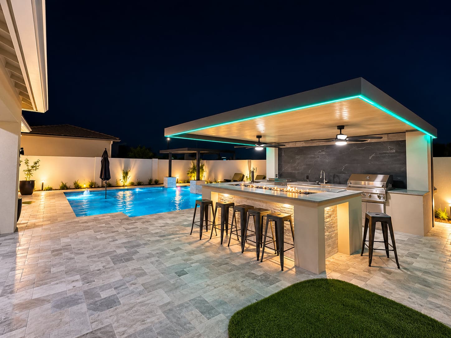 Covered outdoor kitchen and bar beside a lit pool at night, with a teal LED strip along the roof edge.