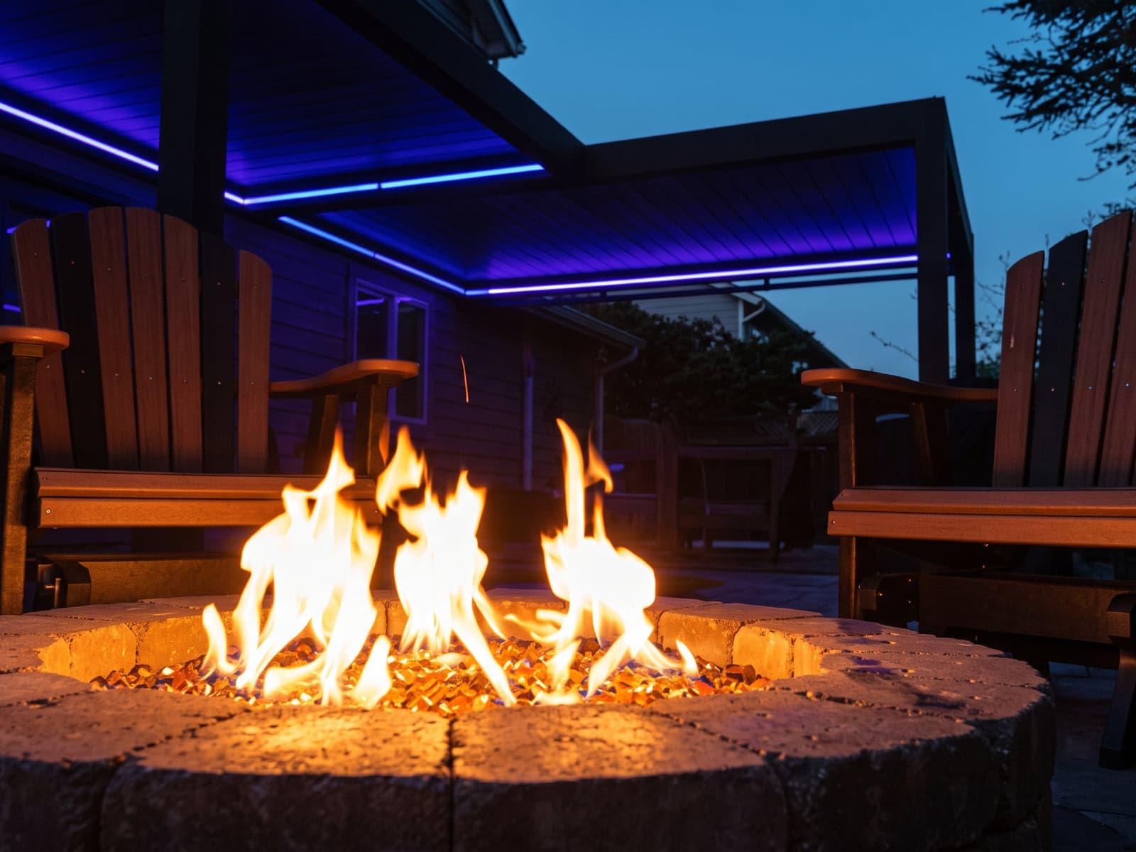 Fire pit with two wooden chairs in front of a covered patio outlined with blue LED strip lights at dusk.